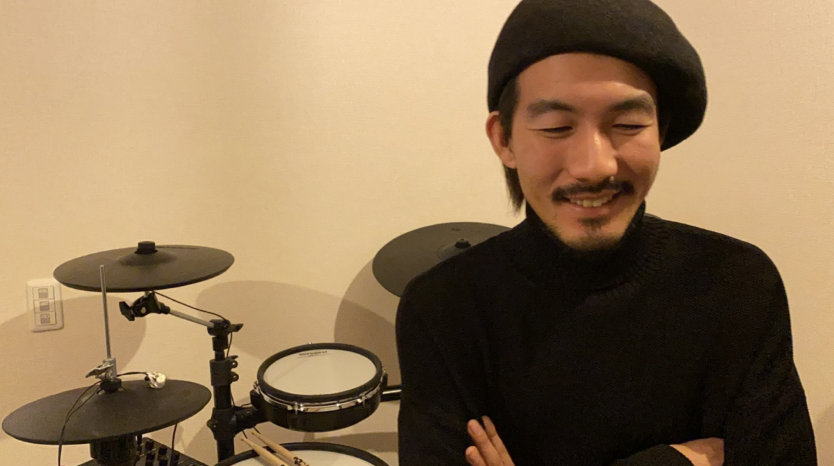 シンドラムの前に座りベレー帽をかぶる神田リョウ