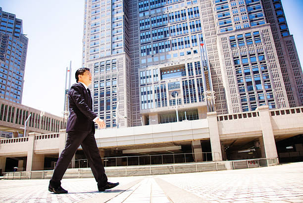 Japanese government worker walking to work in front of Tokyo city hall. Tilted profile view, full length. ei is dressed in a suit and is looking purposefully ahead. Photographed in Shinjuku, Tokyo, Japan.