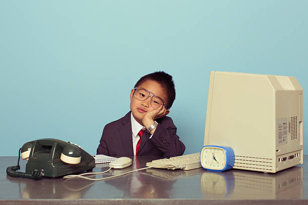 A young Japanese boy dressed as a businessman and executive sits at his desk and is bored from the lack of work. Boy is ready to do good business. Retro styled.